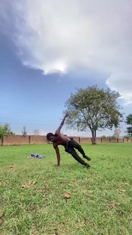 A shirtless, dark-skinned person wearing black leggings is doing an acrobatic flip on a grassy lawn. The person starts in a seated position with their hands and feet on the ground, then pushes off and rotates their body in the air, landing back on their feet. Behind the person is a concrete wall, a few trees, and a blue sky with fluffy white clouds. There's also a blue towel lying on the grass near the wall.