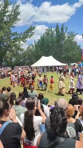 A crowd watches a performance by Indigenous dancers in traditional regalia on a grassy field. A white tent is visible in the background, and trees and a blue sky with clouds complete the outdoor scene. The dancers are performing in a circle, with spectators gathered around them. The performance appears to be a cultural event or festival.