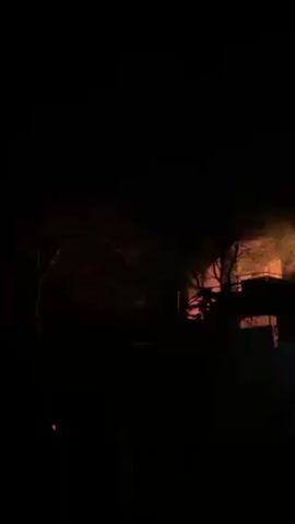 At night, a building is on fire, as seen from a distance. The building appears to be multi-storied, with flames engulfing the top floors. Smoke billows into the dark sky, partially obscuring the view. Bare trees are silhouetted against the fire. Some figures are visible in the lower right corner, but they are too indistinct to identify. The camera remains static throughout the video.