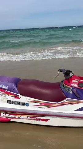 The video shows a close-up of a white Yamaha WaveRunner jet ski parked on a sandy beach. The jet ski has purple and maroon seats, with red and yellow graphics along its side. The registration number 240-51148 is visible. In the background, the turquoise ocean waves are crashing onto the shore under a partly cloudy sky. The camera slowly pans to the right, revealing more of the jet ski and the water.