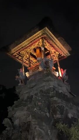 The video shows an illuminated ornate structure at night. The structure appears to be a temple or shrine, with intricate carvings and golden decorations. It is adorned with what look like mythological figures, possibly Garuda, on either side. The camera pans down to reveal a stone inscription that reads "BALE BANJAR UBUD KELOD DESA PAKRAMAN UBUD". The surrounding area is dark, with trees and other foliage visible, suggesting an outdoor setting. The overall impression is one of a sacred or culturally significant site lit up for evening viewing.