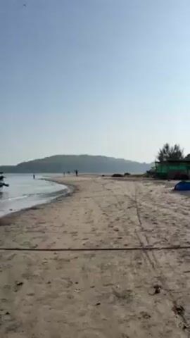 The camera pans across a sandy beach on a sunny day, focusing on several small, wooden boats docked near the shoreline. A green, wooded hill is visible in the background across the water. Several people are walking along the beach in the distance. The water is calm with small waves lapping the shore. There is a rope in the foreground leading to one of the boats.