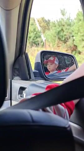A young boy wearing a red and white baseball cap and a red and white striped shirt is visible in the side mirror of a car. His reflection shows him looking forward, with his hand resting on the car door. The camera is positioned inside the car, focusing on the interior, including a black seatbelt and the side panel of the car. The background visible through the mirror and the car window is a blurred, sunny outdoor scene with trees and greenery.