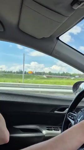 The video shows a first-person perspective from inside a car driving on a road. The driver's hands are visible on the steering wheel, and a watch is on their left wrist. The dashboard and part of the car's interior are visible, including a phone mounted on a holder. Outside the car, a scenic view of a tree-lined road under a partly cloudy blue sky is visible. The car appears to be traveling on a highway with guardrails and sound barriers along the side. There are no other vehicles or people clearly visible in the immediate vicinity of the car. The lighting suggests it is daytime.