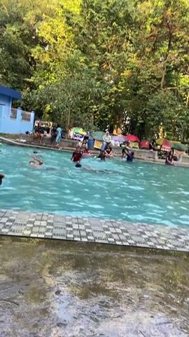 The video shows an outdoor swimming pool filled with people. The pool has blue water and a tiled edge. In the background, there are trees, tents, and some buildings. Several people are in the water, swimming and playing. Some are using inflatable tubes. Others are sitting or standing around the edge of the pool. The overall atmosphere appears to be relaxed and recreational.