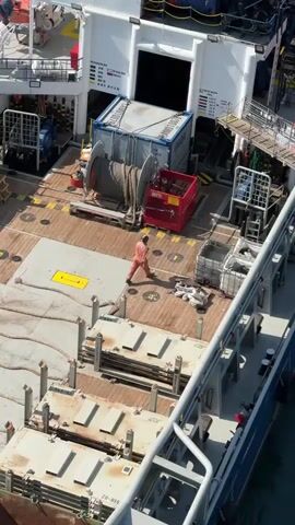 The video shows an aerial view of a ship at sea. The ship is long and narrow, with a white superstructure and a wooden deck. There are several containers and equipment on the deck, including a large winch and a red crane. A person wearing an orange uniform is walking across the deck.

The weather appears to be clear and sunny, with a calm sea. The water is a deep blue color. The ship appears to be in motion, as evidenced by the wake behind it.

The video is shot from another vessel, which is partially visible in the foreground. The other vessel appears to be larger than the ship in the video, and it is also at sea.

The overall impression is one of a working ship at sea. The presence of the worker, the equipment, and the containers suggest that the ship is involved in some kind of industrial activity, possibly related to offshore oil and gas exploration or transportation.

There is some text visible on the ship, including the words "MANIFOLD", "DRY BULK", and "CRANE OIL". There are also some numbers and symbols that are not easily discernible from the video.