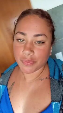 A person with light brown hair in a messy bun and wearing a blue and gray jacket is seen from the chest up, sticking their tongue out. There appears to be a tattoo on the person's chest. The background is blurry, but it seems to be indoors, possibly a bathroom, given the tiled wall visible. The person is looking directly at the camera. Water sounds are audible in the background.