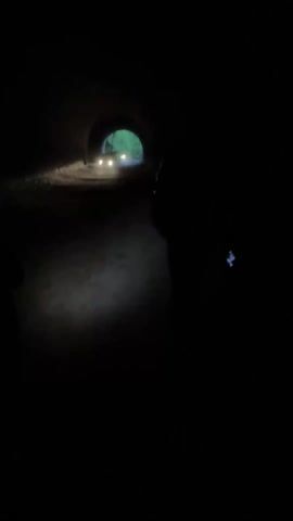 The video depicts a dark tunnel with a bright opening at the far end. Two figures, silhouetted against the darkness, stand inside the tunnel. One figure is more prominent and positioned closer to the center of the frame. A vehicle with its headlights on is seen approaching from the distant tunnel opening, creating a source of bright light amidst the surrounding darkness. The ground appears uneven and covered with loose material, possibly dirt or gravel, illuminated by a light source near the camera.