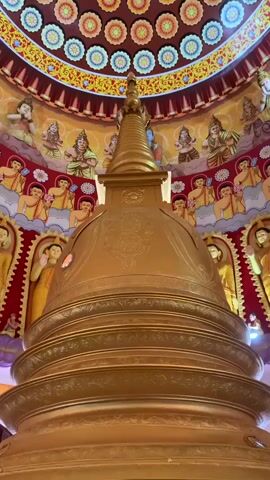A video shows a close-up of a golden stupa inside a temple. The stupa is elaborately decorated, with intricate carvings and a spire at the top. The ceiling of the temple is adorned with a colorful mural depicting floral patterns and figures, possibly deities or monks. The walls also feature murals and sculptures of religious figures. The lighting is warm and inviting, highlighting the golden hues of the stupa and the vibrant colors of the artwork. The camera slowly pans upwards, revealing the grandeur of the temple's interior.
