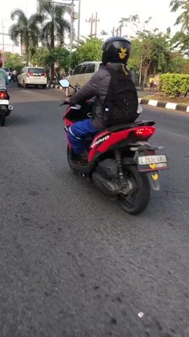The video captures a view from behind a person riding a red and black scooter on a paved road. The rider is wearing a grey jacket, blue pants, and a black helmet with a yellow emblem. They are also carrying a black backpack. The scooter has a license plate that reads "L 393 AB".

In the background, a silver minivan is driving on the same road. Further back, there are trees, palm trees, and some buildings, suggesting an outdoor setting in a somewhat urban or suburban area. The sky appears overcast, indicating it might be a cloudy day or early morning/late afternoon. The lighting suggests it is daytime.

The activity taking place is a person riding a scooter on a road, likely commuting or traveling. The overall impression is a candid shot of everyday life in Candi, East Java, focusing on the movement of the rider and the surrounding environment. No specific emotions are evident due to the focus on the action and the anonymous rider.