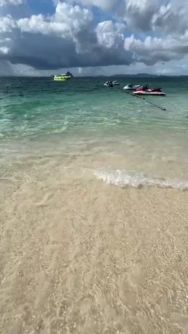 The video captures a serene beach scene with clear turquoise water gently lapping onto a sandy shore. In the distance, a few jet skis are anchored, bobbing on the waves. A larger boat, possibly a ferry, sails further out towards a hazy island on the horizon. The sky above is a mix of dramatic, dark clouds and bright patches of sunlight, suggesting a dynamic weather pattern. The camera angle is low, focusing on the water and sand, giving a sense of immersion in the natural environment. No people are visible in the immediate foreground, and the focus remains on the seascape.