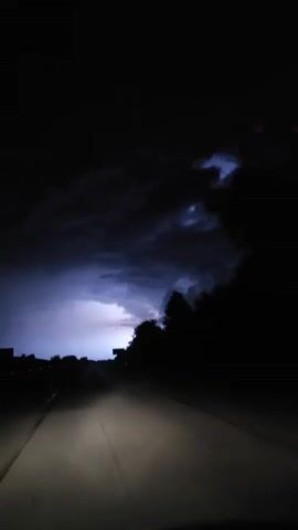 The video displays a first-person perspective from a vehicle driving on a dark road at night. The vehicle's headlights illuminate a segment of the asphalt road directly ahead. Faint white dashed lines are visible on the left side of the illuminated road, with a solid white line on the right. The sky and surrounding environment are completely dark, with only distant, faint red lights visible on the far left horizon. The vehicle is in motion, evidenced by the consistent forward progression of the illuminated road.