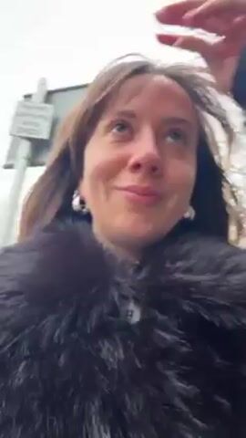 A woman with brown hair is walking outside, smiling at the camera. She is wearing a black fur coat with a button closure. The background is slightly blurred, but trees and a sign are visible. The weather appears overcast.