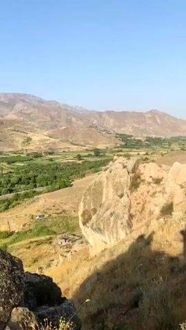 The video begins with a close-up of a large, textured rock in the foreground. The camera then pans left, revealing a vast valley below. The valley is a patchwork of green fields and scattered trees, with a road running through the middle. In the distance, rolling hills and rugged mountains create a dramatic backdrop against a clear blue sky. As the camera continues to pan, a small village comes into view, nestled amongst the trees. Houses with red-tiled roofs are visible, along with a few vehicles. The sun is shining, casting long shadows and illuminating the landscape. The overall impression is one of a peaceful rural setting, bathed in the warm light of day.
