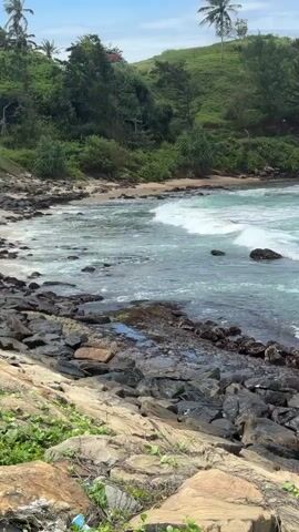 A static, medium shot captures a tropical coastline with a rocky shore in the foreground. Waves crash against dark, jagged rocks, creating white foam that spreads into the turquoise water. Beyond the water, a sandy cove is visible, backed by a steep, green hill covered in lush vegetation, including trees and palm trees. The sky is clear blue with a few scattered white clouds. The camera is angled slightly upwards, emphasizing the height of the hill and the expanse of the ocean. No people or man-made structures are clearly visible in the frame, apart from what may be a distant fence or utility pole on the hillside. The overall atmosphere is serene and natural.