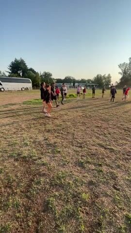 The video shows a group of people in a grassy field under a bright sun. The people appear to be engaged in some form of outdoor activity or training. Some individuals are performing kicks and stretches, while others are standing or walking in formation. The environment is open with some trees and buildings in the background. The sun is low in the sky, casting long shadows, suggesting it is either early morning or late afternoon. There is a bus visible on the left side of the frame. The overall scene depicts an organized group exercise session in an outdoor setting.