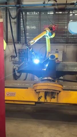 A robotic arm is welding a metal beam. The robotic arm is yellow with a red end effector, and it is positioned above the metal beam. The welding process is creating a bright blue light and sparks. The metal beam is on a yellow conveyor belt system. The background shows a factory setting with windows and machinery.