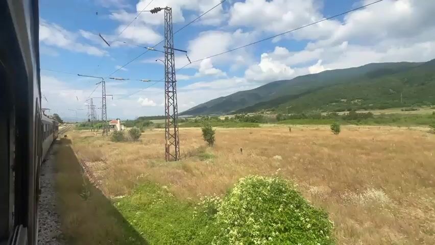 The video captures a view from the window of a moving train. The train's side is visible on the left. The landscape outside consists of dry, golden grass fields and some scattered bushes and trees. In the distance, green, tree-covered mountains form the background under a blue sky with white clouds. Tall electricity pylons with wires are prominent in the middle ground. A small building is visible near the train tracks in the distance. The train appears to be moving along a set of tracks that curve towards the left side of the frame. The overall scene suggests a rural or countryside setting during the daytime, possibly in the summer or late spring due to the dry grass.