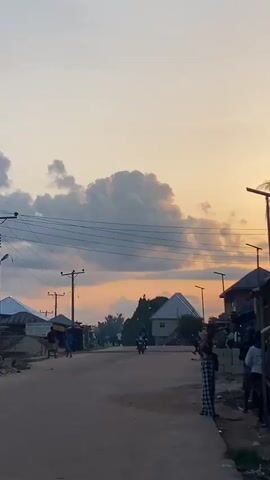 The video shows a street scene at dusk. A dirt road stretches into the distance, lined with buildings and trees. A few people are visible on the street, some standing and talking, others walking or riding motorcycles. The sky is filled with dramatic clouds, illuminated by the setting sun, casting a warm glow over the scene. Power lines crisscross the sky, and streetlights are visible on poles. The overall impression is of a quiet, communal evening in a small town.