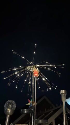 The video shows an outdoor setting at night. A decorative light feature, resembling a burst of fireworks, is prominently displayed, with numerous thin, flexible strands radiating outwards, each tipped with a small, bright LED. At the center, there is a small orange flower detail. The light feature is mounted on a set of metal poles. Below it is a small, colorful pinwheel. The background is dark, with the silhouette of a building visible along the horizon. To the sides of the main light feature are two additional light fixtures, one with a clear, textured glass sphere and another that is mostly out of frame. The overall atmosphere is calm, with the focus on the decorative lights illuminating the night.