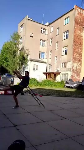 In this video, a man is performing a juggling act with a ladder in an outdoor courtyard. He is wearing a dark t-shirt and shorts, and appears to be practicing or performing in front of an audience. The setting is urban, with a tall, light-colored apartment building in the background. The building has several windows and balconies, and some of the brickwork on the right side is exposed and appears weathered. There are trees and bushes around the building, suggesting it is a residential area. In the foreground, the courtyard is paved with concrete. Several objects are scattered on the ground, including what appear to be other juggling props or equipment, a red cooler, and a car. A second person is partially visible in the background, near a car, and another person is on a balcony in the building. The sky is clear and blue, indicating a sunny day. The man's movements are fluid and coordinated, suggesting he is skilled at juggling. The overall atmosphere is casual and energetic, with the focus on the man's performance. There is no visible text in the video. The time of day appears to be daytime, possibly late morning or early afternoon, given the bright sunlight.