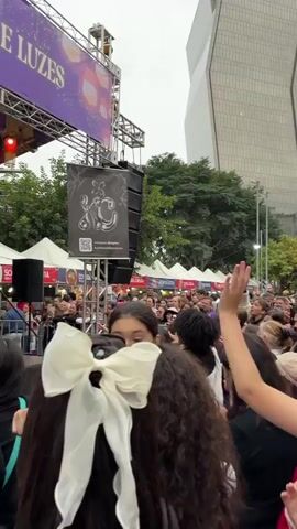 A crowd of people is gathered in front of a stage, watching a live performance. On the stage, there are two dancers and a blonde singer. The performers are wearing black clothing. The singer is holding a microphone and dancing. The backdrop is a banner with a name and logo on it. Above the stage are hanging colorful lanterns. A tall office building is visible in the background.