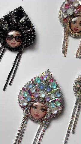 The video shows a top-down view of four handmade brooches arranged on a white surface. Each brooch is in the shape of a heart and features a miniature painted portrait of a woman's face at the bottom. The brooches are adorned with numerous rhinestones and jewels in various colors, forming elaborate headdresses. The top left brooch is black and features sunglasses on the portrait. The other three brooches have colorful headdresses with varying shades of pink, blue, and iridescent stones. All brooches have dangling chains of rhinestones extending from the sides. The lighting is bright, highlighting the sparkle of the jewels. The camera pans slowly across the brooches, providing a detailed view of each one. There are no people or active interactions depicted, only the static display of the brooches.