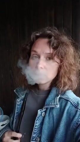 A young woman with curly brown hair and wearing a denim jacket exhales a plume of smoke in this close-up, low-angle shot. She is holding a dark-colored hookah hose in her left hand. The background appears to be a dark, wooden wall, suggesting an indoor or covered outdoor setting. The lighting is dim, creating a dramatic effect with the smoke obscuring part of her face. She seems to be looking directly at the camera, with a neutral expression. The smoke is the most dynamic element in the frame, swirling and dissipating around her head. There is no visible text in the image. The scene is dark and moody.