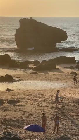 A beach scene at sunset with the sun low on the horizon casting a warm glow. Several people are present on the sandy beach and in the ocean. In the foreground, two women are standing near a blue umbrella. Further back, two young men are playing with a frisbee. In the mid-ground, a group of young men are playing soccer with a ball. In the background, a large rock formation is visible in the ocean, with waves crashing against it. Several other people are visible in the water in the distance. The overall atmosphere is relaxed and leisurely.