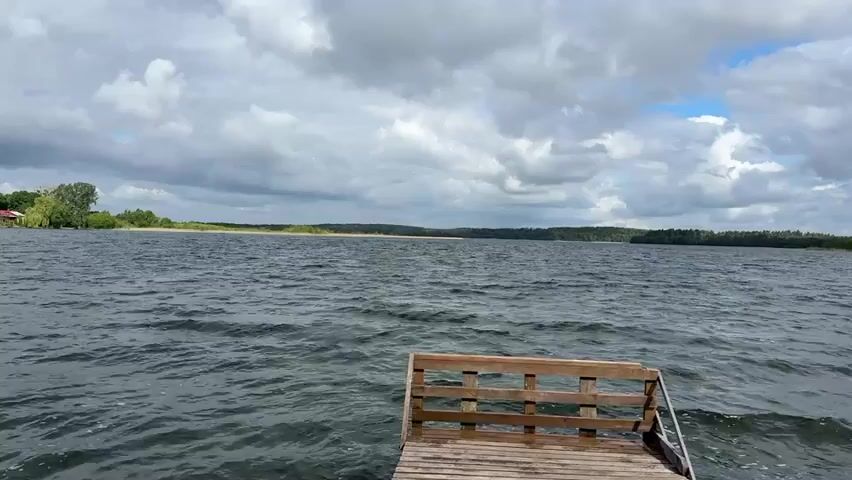 The video presents a still shot of a choppy lake under a cloudy sky, viewed from a wooden dock. The dock has a small, wooden railing at its edge. In the distance, there is a low shoreline covered with trees and vegetation. The sky is filled with grey and white clouds. The water is dark and rippled, indicating some wind activity. No people are visible in the shot.