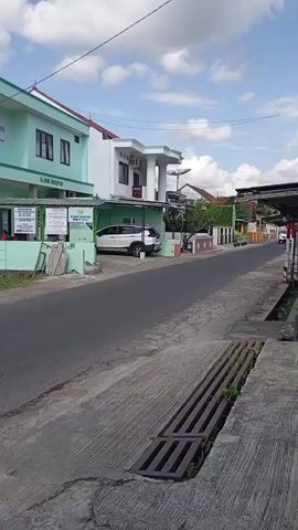 A vertical, wide shot shows a quiet street in a neighborhood. On the left is a light green building with white trim and windows, labeled "KLINIK MERPATI". A white SUV is parked in front of it. An ambulance with flashing lights drives past on the road, and a motorcycle is seen in the distance on the right. The street is paved, with a concrete drain cover in the foreground. The sky is partly cloudy with a bright sun.