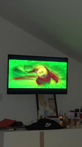 The video shows a television screen displaying scenes from the animated movie Kung Fu Panda 4. In the first scene, the screen is filled with bright green and yellow abstract imagery. In the second scene, a large blue creature with horns is visible, set against a blue, mountainous landscape. The third scene returns to the green and yellow abstract imagery. The fourth scene features Po, the panda protagonist, wearing a conical hat and a green robe, surrounded by a green, glowing effect. The fifth scene shows the blue creature with horns again, with a more detailed view of its face. The television is mounted on a wall above a dresser. On the dresser, there is a framed picture of a man and a woman, several small figurines, and various bottles and boxes. The room appears to be dimly lit.