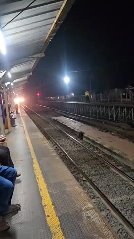 The video captures a night scene at a train station platform in Tenjo, Indonesia. A train is seen approaching from the distance, its bright headlights illuminating the tracks ahead. Multiple train tracks run through the center of the frame.

On the left side, under a covered platform, several people are waiting. Some are seated, while others appear to be standing, looking towards the approaching train. One individual in blue pants and sandals is clearly visible in the foreground, seated on the platform.

Across the tracks, on another platform to the right, a station staff member wearing a high-visibility vest can be seen standing. A red signal light is also visible further down the track.

As the train draws nearer, a train horn sounds, and a voice announces something over the station's public address system. The scene is lit by various artificial lights, including overhead lights on the platform and the powerful headlights of the train, creating a typical nighttime atmosphere at a busy train station.