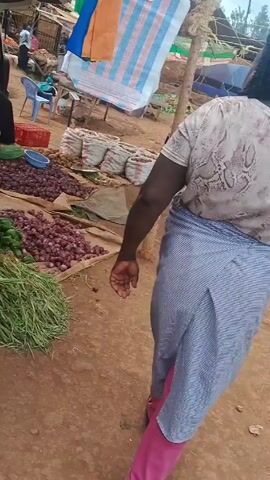 The video shows a bustling market scene. A person with a striped skirt is standing near a display of tomatoes, green peppers, onions, and herbs arranged on a brown surface. Another person wearing a green patterned skirt walks in the background. A variety of vegetables, including potatoes and onions, are displayed in sacks and on mats.  A person scans a basket filled with tomatoes and herbs using a smartphone. A car is visible in the background. The ground is reddish-brown.