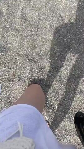 The video shows a first-person perspective of someone walking on a pebbly beach. The person is wearing a white dress and flip-flops. Their shadow is cast on the ground. The lighting suggests it is daytime and sunny. The camera is pointed downwards, focusing on the feet and the ground. There is a white crocheted bag visible in the lower left corner of the frame. The scene is static with the only movement being the walking motion of the person.