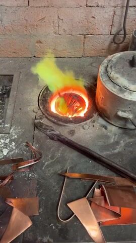 The video captures a close-up view of a metal melting operation taking place in what appears to be a workshop or industrial setting, likely in Saint-Rémy-lès-Chevreuse, France.

In the foreground and center, a small, round crucible is embedded within a furnace, glowing intensely with a bright orange and red heat. Inside the crucible, various scraps of reddish-brown metal, strongly resembling copper, are visible and appear to be in the process of melting. A prominent yellow-green flame or smoke billows upwards from the crucible, indicating extreme temperatures and active combustion or the burning off of substances.

To the immediate right of the furnace, a larger, cylindrical metal pot or crucible with a lid is partially visible, also appearing well-used and possibly hot. Resting on the concrete floor in front of the furnace is a long, dark metal tool, possibly tongs or a stirring rod. More flat, reddish-brown metal scraps, consistent with copper, are scattered on the floor to the left and bottom right of the main melting area.

The background features a rough, reddish-brown brick wall, suggesting a sturdy and heat-resistant construction for the workspace. The overall scene is one of intense heat and metal transformation. No people are visible, nor is any discernible text present in the frame. The audio features a loud, continuous roaring sound, characteristic of a powerful furnace burner in operation. The time of day and weather are not discernible, as the scene is indoors.