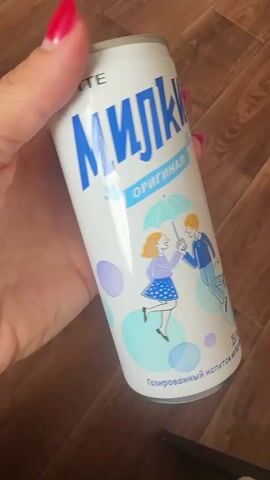 A hand with red nail polish is holding a white can of a drink with blue lettering. The can has a cartoon image of a boy and a girl holding an umbrella together. The background is a wooden floor.