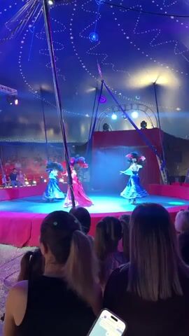 The video shows a circus performance inside a tent. The tent ceiling is decorated with blue fabric and illuminated with star-like lights. A circular stage is lit with red and blue spotlights. Three performers in elaborate costumes, featuring puffy sleeves and large headdresses, are dancing on the stage. Two of the performers are wearing blue and silver dresses, while the third is wearing a red and gold dress. The performer in the red and gold dress is positioned in the center of the stage and appears to be leading the dance. The audience, consisting of several people, is visible in the foreground, with their backs to the camera, watching the performance. The overall atmosphere is vibrant and festive, with smoke effects adding to the dramatic presentation.