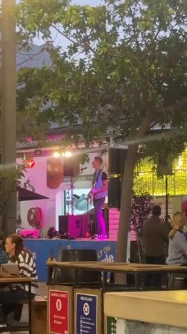 A musician plays guitar on a small outdoor stage in front of a building. He wears a blue denim vest, dark shirt, and jeans. The stage is decorated with colorful graphics. Stage lights illuminate him in pink and white. To the sides of the stage are trees. Several people sit at picnic tables in front of the stage, appearing to watch the performance. A trash and recycling bin are visible near one of the tables. The sky is dusky, suggesting early evening.