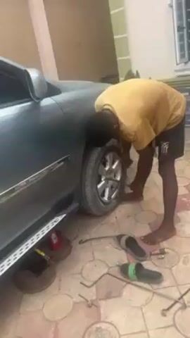 A man is changing a tire on a car in what appears to be a driveway or patio. He is wearing a yellow shirt and black shorts with a graphic and is barefoot. The car is a dark color, possibly gray or blue, and is raised with a jack. Several tools, including lug wrenches and a spare tire, are on the ground around him. The ground is paved with patterned tiles, and a building with a window is visible in the background. The man is focused on the lug nuts of the tire.