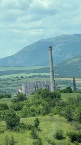A wide shot captures a view of a large, industrial power plant set against a backdrop of rolling hills and mountains. The power plant features a prominent, tall, slender smokestack and a long, rectangular building with numerous vertical architectural elements. The surrounding landscape is characterized by lush green fields and scattered trees, suggesting a rural or semi-rural environment. The sky is partly cloudy, with patches of blue visible. The video appears to be filmed from a moving vehicle, as indicated by the slight blur and a lens flare that briefly appears in the lower-left portion of the frame. No people or discernible activity are visible within the frame.