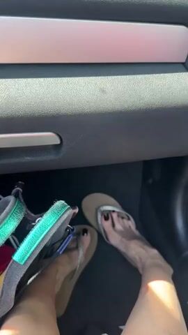 The video shows a person's feet wearing flip-flops inside a car, transitioning to a view through the car's windshield while driving on a highway. The initial shot focuses on the lower part of the car's dashboard and the person's legs, highlighting the flip-flops they are wearing. One pair is a tan color with a silver strap, the other is more athletic-style, with teal and black straps. The person has dark nail polish on their toes. The video then shifts to a view from the driver's perspective, showing the highway with other vehicles in motion on a sunny day. A barrier lines one side of the road, covered in greenery, and the sky is clear and blue. The movement indicates the car is in transit.