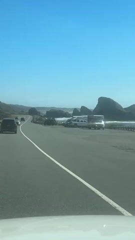 A vehicle is traveling along a highway. The highway runs parallel to a rocky coastline with the ocean visible in the background. Several vehicles are parked on the shoulder of the highway, and some people appear to be near them. The sky is clear and blue.