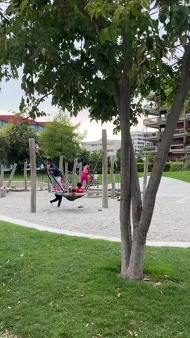 A man and a woman are supervising three young children on a playground. The children are on a hammock-like swing, with one child in the hammock and two others standing and holding onto the ropes, appearing to push the swing. The playground has a surface of white gravel and features wooden poles and rope structures. The surrounding environment includes a grassy area, trees, and modern apartment buildings in the background. The sky is overcast.