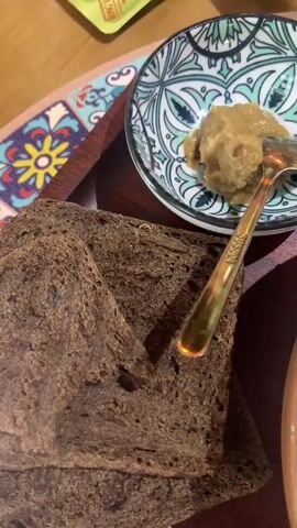 A close-up shot shows two slices of dark bread next to a small bowl containing a pale, chunky substance and a spoon. To the right of the bread and bowl, a large ceramic bowl filled with a light-colored, creamy soup with visible green herbs and red onion slices. A spoon is partially submerged in the soup. The items are resting on a wooden tray, which is on top of a placemat with a colorful pattern.