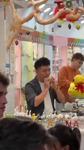 The video shows a man in a black blazer blowing into a small, yellow ocarina-like instrument. He stands behind a display case with small ceramic animals and the word "Iwanjocarina" on it. To his right, another man in an orange shirt stands looking in the same direction. In the foreground, the heads of other individuals are visible, suggesting a crowded environment. The background reveals a colorful store interior, with shelves displaying various items and decorative elements like a branch with ornaments hanging from the ceiling.