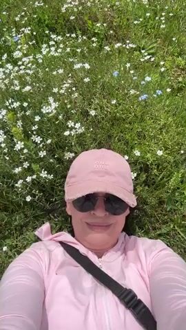 A woman lies on her back in a field of white and blue wildflowers, wearing a pink baseball cap and sunglasses. She is also wearing a pink hooded jacket with a black strap across her chest. The camera is held at an angle to capture her face and the flowers around her. The sun is shining, and the overall scene suggests a relaxed outdoor setting.