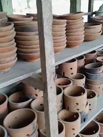 The video shows a series of shelves filled with pottery and bags of soil. The top shelf has large, shallow bowls and white concrete decorations. The lower shelves are filled with terracotta pots, some of which have holes in the sides. The shelves are made of unfinished wood. The environment appears to be a shop or storage area.