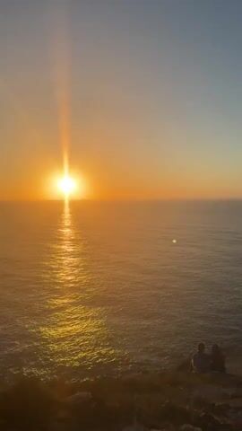 The video shows a sunset over the ocean. There are several people on a cliff overlooking the water. The sun is setting on the left side of the frame, casting a warm glow over the scene. The water is calm, and the sky is a gradient of orange and blue. Some people are sitting on the cliff, while others are standing and talking. The overall atmosphere is peaceful and serene.