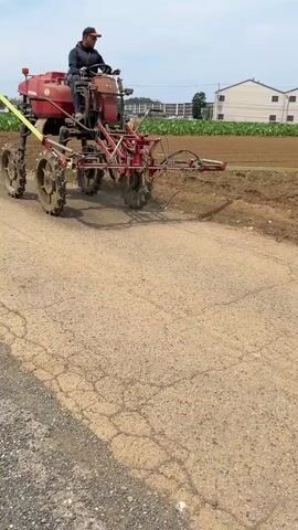 A person is operating a red agricultural machine with large wheels and a spray tank on a dirt road. The machine has a red frame with various mechanical components. The person is wearing a dark jacket, blue pants, a hat, and gloves, and is seated, controlling the machine with a steering wheel. The machine is spraying something onto a tilled field on the right side of the road. The field is brown and appears to be recently worked. In the background, there are some buildings and trees under a cloudy sky. A yellow strap is attached to the front of the machine and extends out of frame to the left. The road surface is cracked and dry. The machine's wheels are covered in mud.